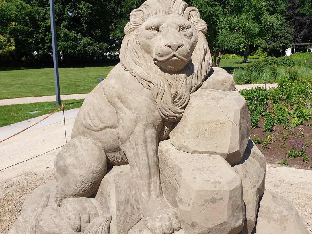 Eine detailgetreue Sandstatue eines majestätischen Löwen, umgeben von grünen Bäumen und Pflanzen.