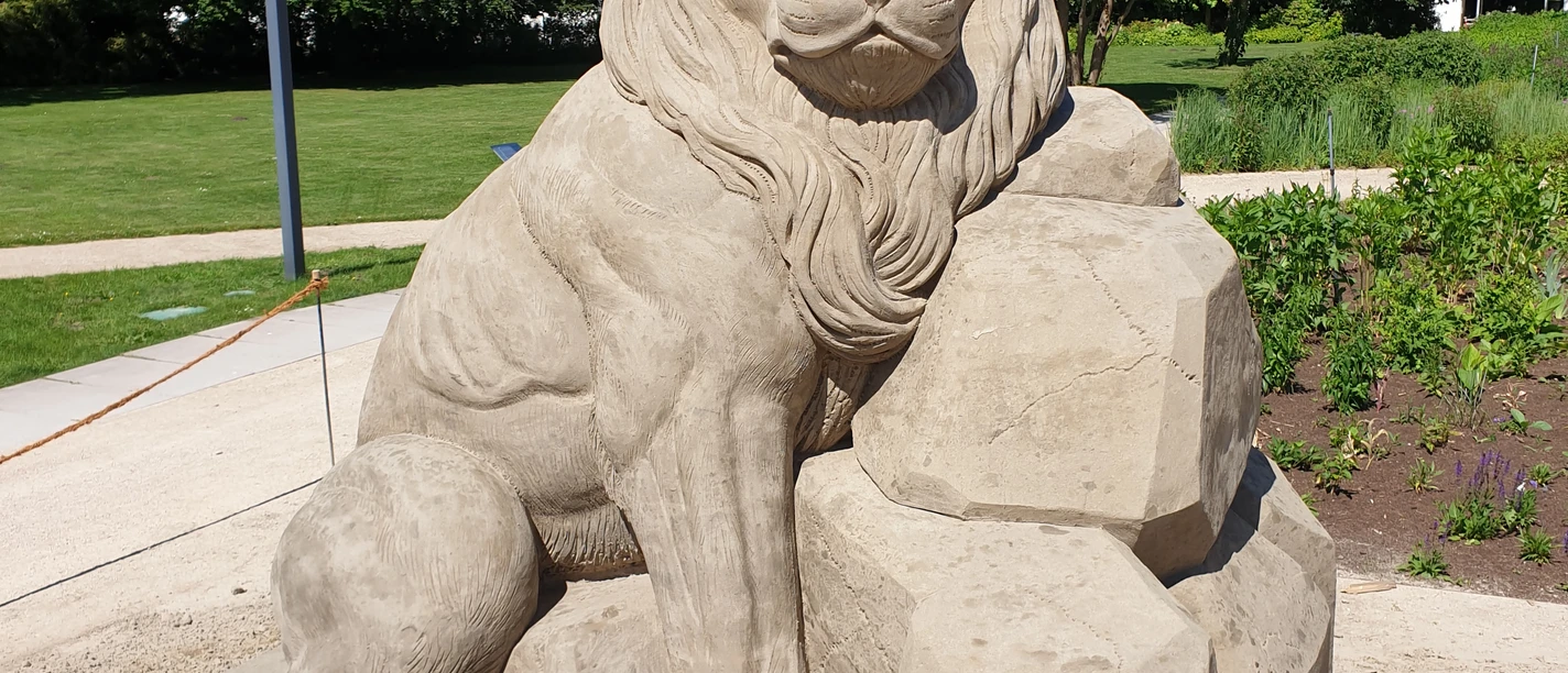 Eine detailgetreue Sandstatue eines majestätischen Löwen, umgeben von grünen Bäumen und Pflanzen.