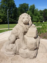 20220607_113932.jpg Eine detailgetreue Sandstatue eines majestätischen Löwen, umgeben von grünen Bäumen und Pflanzen.
