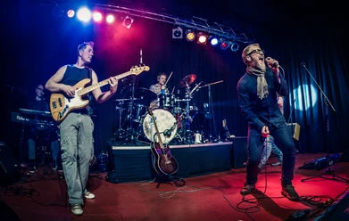 Eine Band rockt eine Bühne mit E-Gitarre, Schlagzeug und ausdrucksstarkem Sänger in actionreicher Beleuchtung.