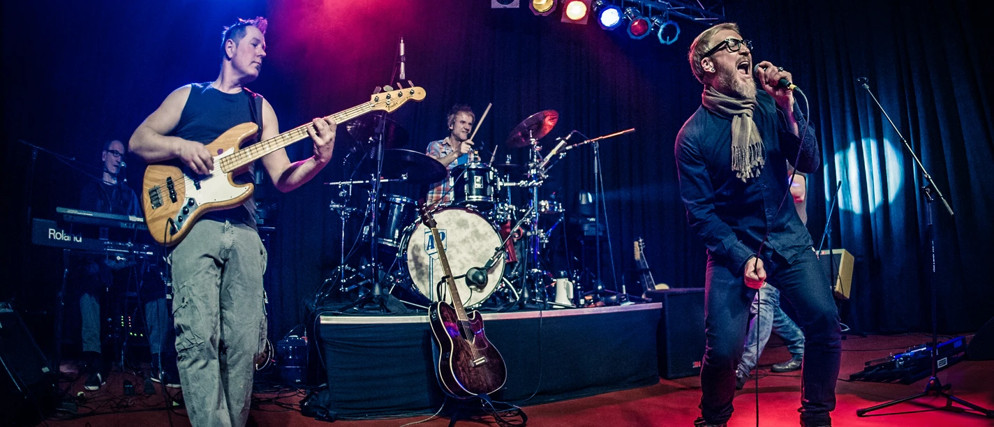 AP Rene 1.jpg Eine Band rockt eine Bühne mit E-Gitarre, Schlagzeug und ausdrucksstarkem Sänger in actionreicher Beleuchtung.