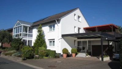 Ferienwohnung Brigitte u. Wolfgang Waldow.jpg Zweistöckiges Haus mit weißer Fassade, darüber ein blauer Himmel; links Wintergarten.