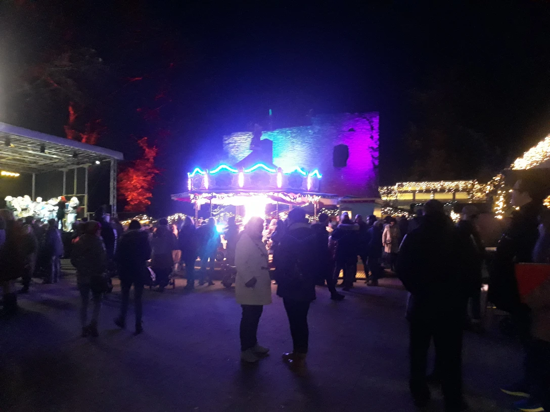 20191130_174050.jpg Nachtszene eines Weihnachtsmarktes mit farbenfroher Beleuchtung, Menschenmenge und erleuchteten Ständen.