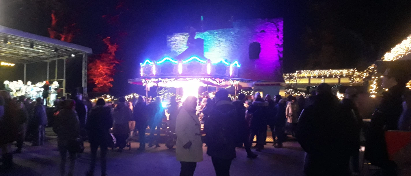 20191130_174050.jpg Nachtszene eines Weihnachtsmarktes mit farbenfroher Beleuchtung, Menschenmenge und erleuchteten Ständen.