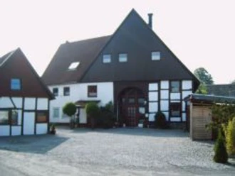 Fachwerkhaus mit dunklem Satteldach, gepflasterter Hof, umgeben von grünen Büschen und Bäumen.
