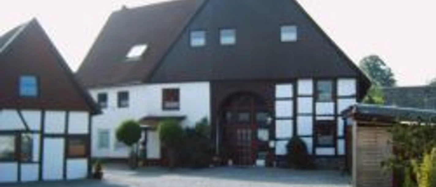 Fachwerkhaus mit dunklem Satteldach, gepflasterter Hof, umgeben von grünen Büschen und Bäumen.