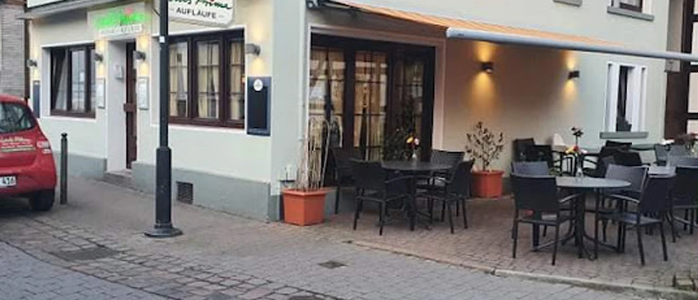 Louis Prima.jpg Pizzeria mit Außenterrasse und roten Dachziegeln in ruhiger Seitenstraße, Laterne und Auto daneben.