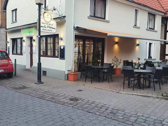 Louis Prima.jpg Pizzeria mit Außenterrasse und roten Dachziegeln in ruhiger Seitenstraße, Laterne und Auto daneben.