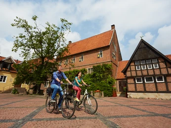 Route 9_Welfen_Westerholtscher Burgmannshof Kornbrennerei Berentzen in Haselünne - CR Emsland Tourismus GmbH (8).jpg Zwei Radfahrende auf einem gepflasterten Platz vor historischen Backsteinbauten in Haselünne.