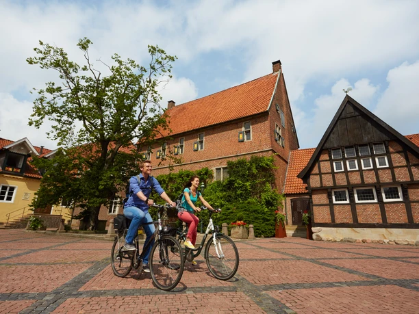 Route 9_Welfen_Westerholtscher Burgmannshof Kornbrennerei Berentzen in Haselünne - CR Emsland Tourismus GmbH (8).jpg Zwei Radfahrende auf einem gepflasterten Platz vor historischen Backsteinbauten in Haselünne.