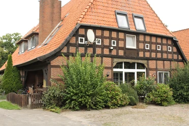 Vakwerkhuis met rode dakpannen en grote ramen, omringd door weelderige vegetatie en struiken.