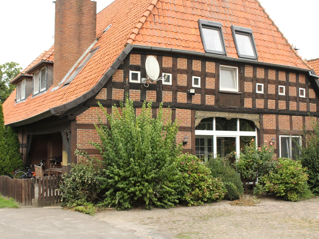 Zum Fletthof Fachwerkhaus mit roten Dachziegeln und großen Fenstern, umgeben von üppiger Vegetation und Sträuchern.Half-timbered house with red roof tiles and large windows, surrounded by lush vegetation and shrubs.Bindingsværkshus med røde tagsten og store vinduer, omgivet af frodig vegetation og buske.Vakwerkhuis met rode dakpannen en grote ramen, omringd door weelderige vegetatie en struiken.