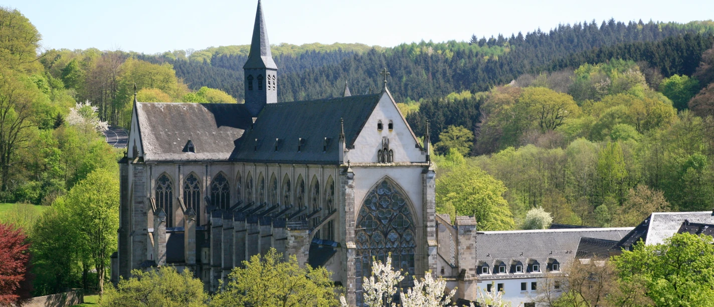 Altenberger Dom Eine gotische Abtei inmitten grüner Wälder und blühender Bäume unter klarem, blauem Himmel.