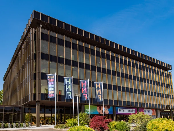 Ein modernes, mehrstöckiges Gebäude mit farbigen Fahnen und Glasfassade unter blauem Himmel.