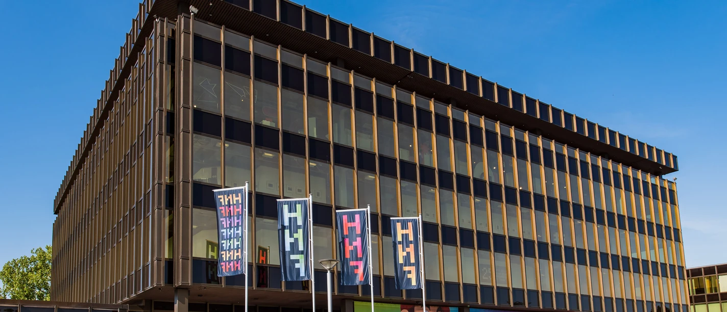 Fahnen_2022_HNF_5.jpg Ein modernes, mehrstöckiges Gebäude mit farbigen Fahnen und Glasfassade unter blauem Himmel.