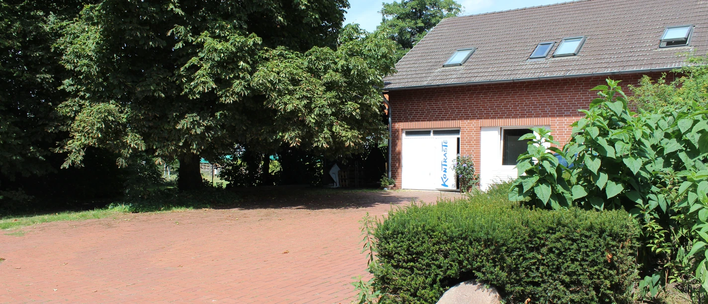 Bakstenen gebouw met zadeldak en dakramen, omgeven door dichte begroeiing en straatstenen.