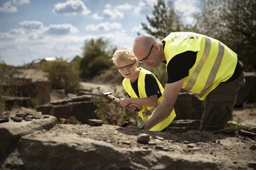 Fossiliensuche <p>Ein Erwachsener und ein Kind in gelben Westen erforschen gemeinsam Gesteine im Freien.</p>