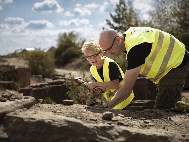 Fossiliensuche <p>Ein Erwachsener und ein Kind in gelben Westen erforschen gemeinsam Gesteine im Freien.</p>