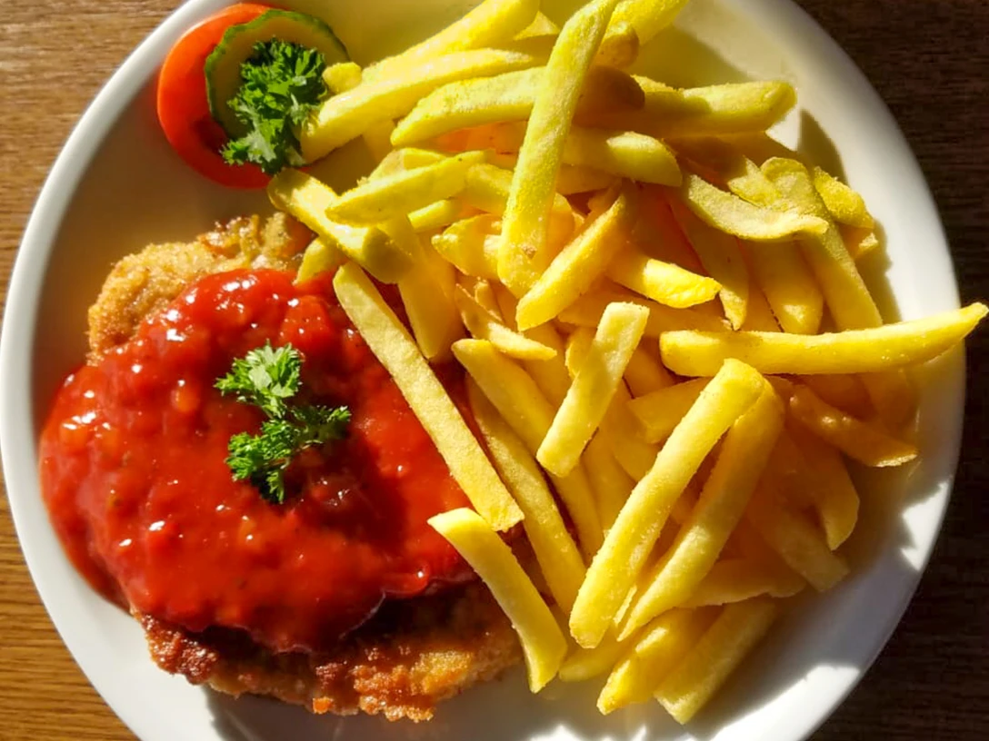 Schnitzel mit roter Soße und grüner Garnitur, serviert mit knusprigen Pommes auf weißem Teller.
