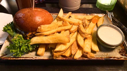 Ein Cheeseburger mit grünen Salatblättern, serviert mit dicken Pommes und weißem Dip auf brauner Platte.