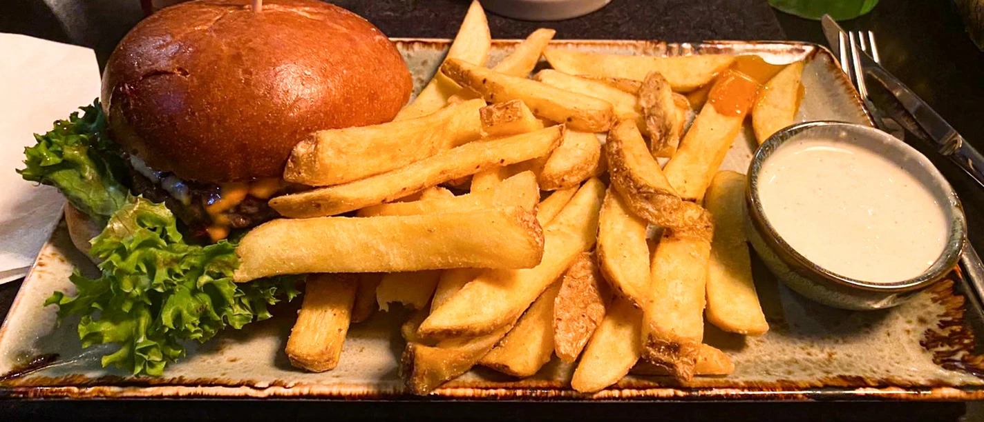 Ein Cheeseburger mit grünen Salatblättern, serviert mit dicken Pommes und weißem Dip auf brauner Platte.