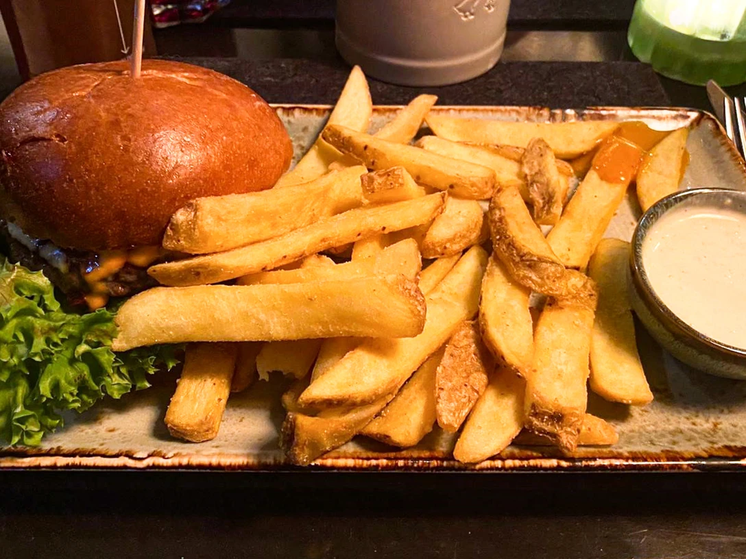 Hamburger-Pommes_CCBYSA_Teutoburger Wald_Stadt Schloss Holte-Stukenbrock-8.jpg Ein Cheeseburger mit grünen Salatblättern, serviert mit dicken Pommes und weißem Dip auf brauner Platte.