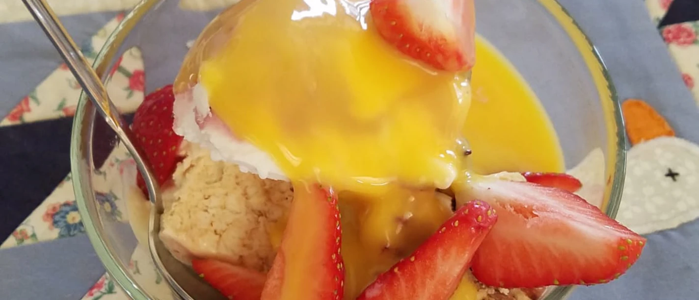 Glas mit Vanilleeis, Erdbeerscheiben und gelber Soße auf einer Tischdecke platziert.