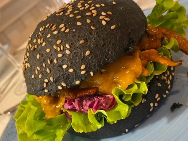 Burger schwarz_CCBYSA_Teutoburger Wald_Stadt Schloss Holte-Stukenbrock-13.jpg Schwarzer Burger mit Sesam, gefüllt mit Salat, rotem Kraut, Käse und gegrilltem Hähnchen auf Teller.