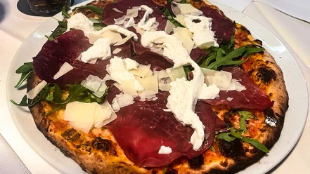 Eine Steinofenpizza mit Bresaola, Rucola, Büffelmozzarella und Parmesanschnipseln auf einem Teller.