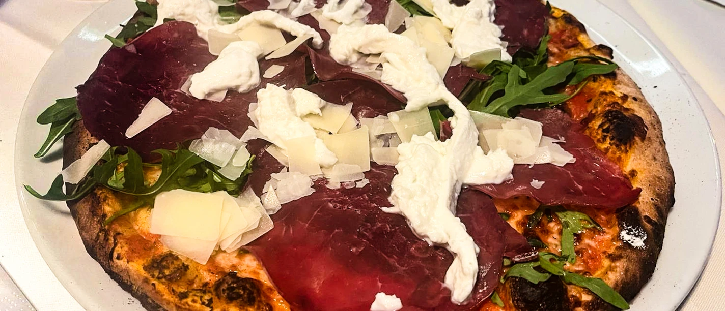 Pizza2_CCBYSA_Teutoburger Wald_Stadt Schloss Holte-Stukenbrock-11.jpg Eine Steinofenpizza mit Bresaola, Rucola, Büffelmozzarella und Parmesanschnipseln auf einem Teller.
