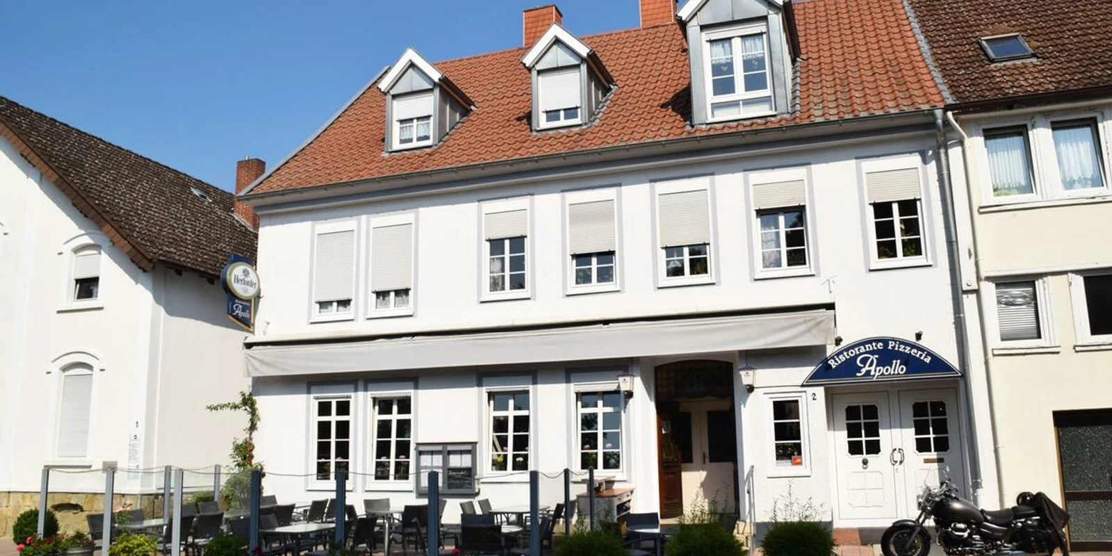 Restaurant Apollo.JPG Das Bild zeigt ein großes Restaurantgebäude mit roten Dachziegeln und einer Außenterrasse bei Sonnenschein.