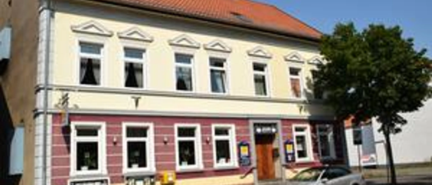 Zweistöckiges Gebäude mit gelber Fassade, roten Akzenten und geparktem silbernen Auto davor.