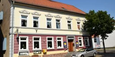 Zweistöckiges Gebäude mit gelber Fassade, roten Akzenten und geparktem silbernen Auto davor.