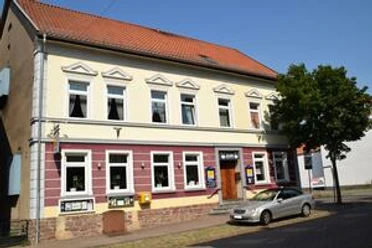 Bei Heini Ansicht.JPG Zweistöckiges Gebäude mit gelber Fassade, roten Akzenten und geparktem silbernen Auto davor.