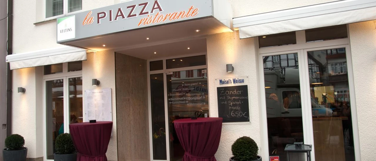 La Piazza Ristorante.JPEG Eingangsbereich zum Restaurant mit Vordach und zwei Rundtischen, dekoriert mit roten Tüchern.