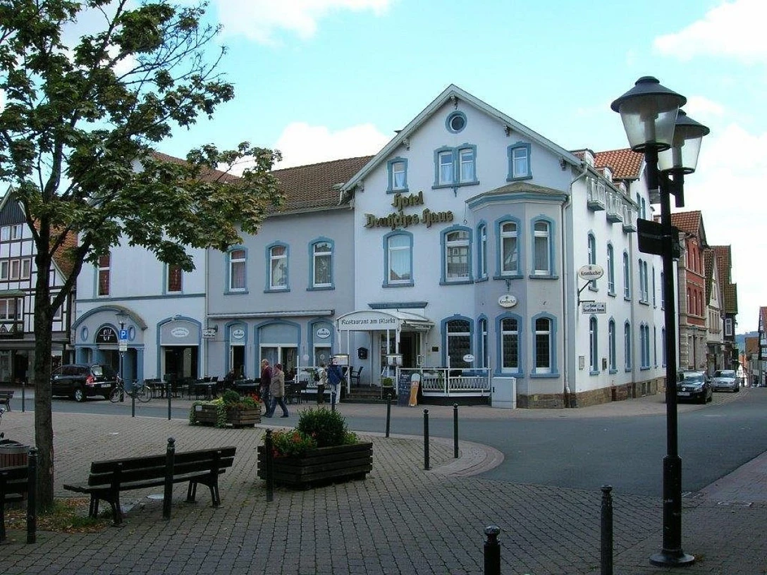 Hotel Deutsches Haus.jpg Hotel Deutsches Haus an einer Straßenecke mit weißer Fassade, umgeben von Bäumen und Laternen.