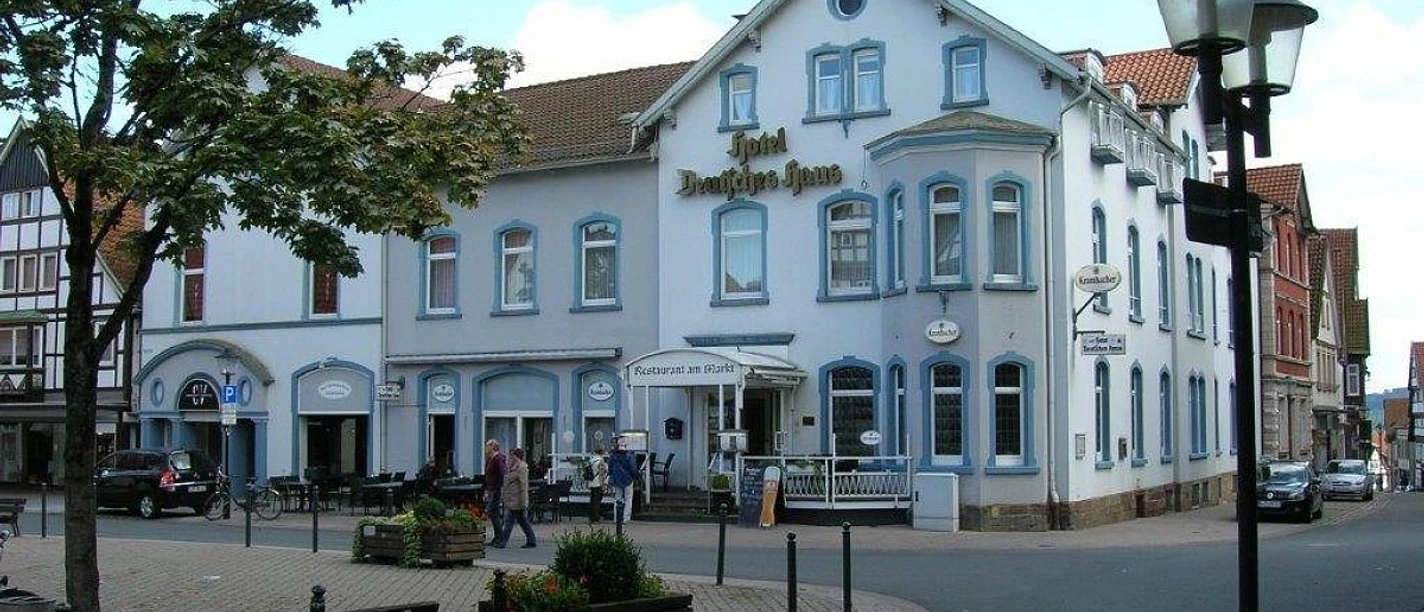 Hotel Deutsches Haus.jpg Hotel Deutsches Haus an einer Straßenecke mit weißer Fassade, umgeben von Bäumen und Laternen.
