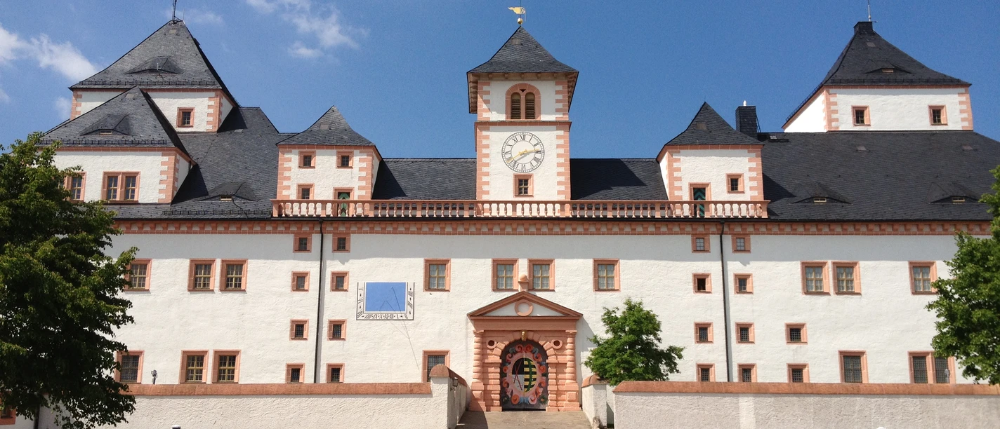 Schloss mit weißen Mauern, roten Akzenten und Schieferdächern bei strahlend blauem Himmel.