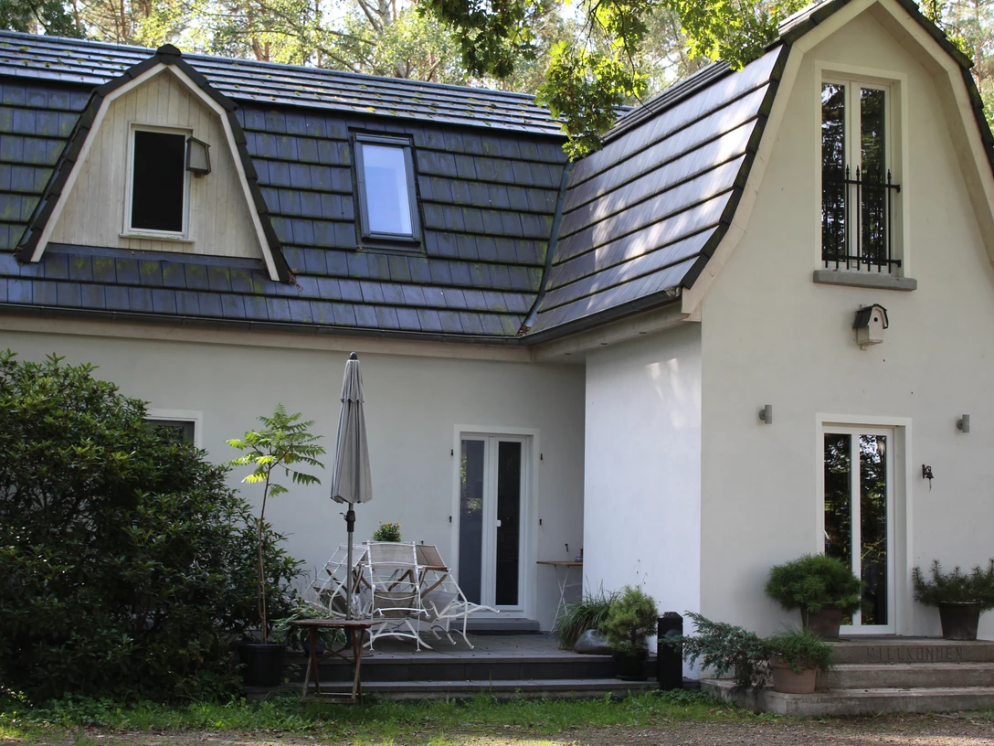 Altes Waldhäuschen Ein kleines weißes Haus mit blauer Dachdeckung, umgeben von Bäumen und einem Terrassenbereich.