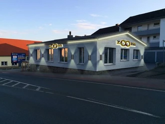 zoom.jpg Ein einstöckiges Gebäude mit „zoom“ Beschriftung, das von warmem Kunstlicht bestrahlt wird.
