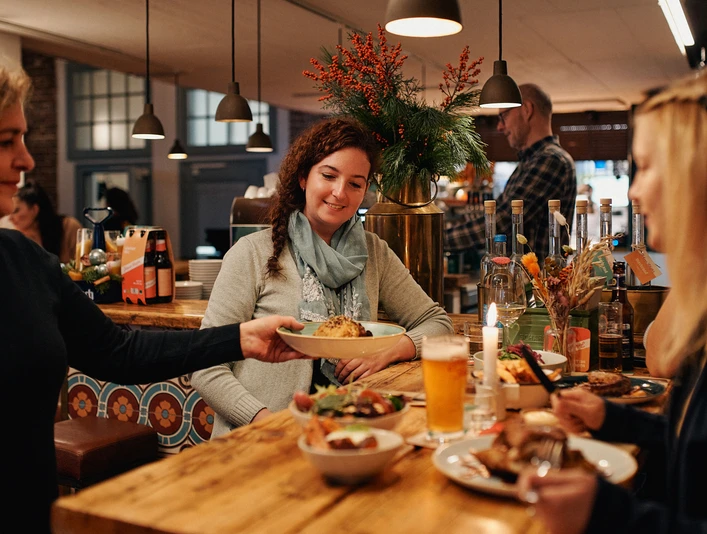 Brauhaus Johann Schäfer Zwei Frauen sitzen an einem naturbelassenem Holztisch, in einem gemütlich warm beleuchteten Restaurant. Eine Kellnerin serviert einen Teller.Two women are sitting at a natural wooden table in a cozy, warmly lit restaurant. A waitress is serving a plate.