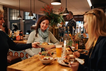 Johann Schäfer Brewery Two women are sitting at a natural wooden table in a cozy, warmly lit restaurant. A waitress is serving a plate.