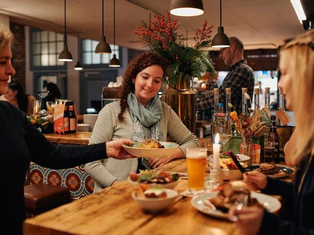 Johann Schäfer Brewery Two women are sitting at a natural wooden table in a cozy, warmly lit restaurant. A waitress is serving a plate.