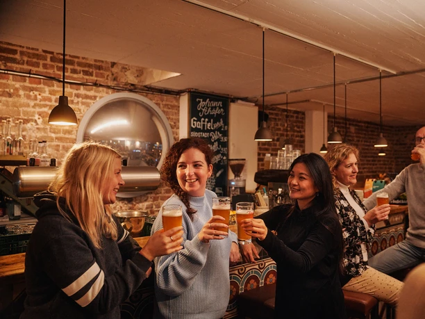 Brauhaus Johann Schäfer Drei Frauen stehen an der Bar des Restaurants und stoßen mit einem Bier an.