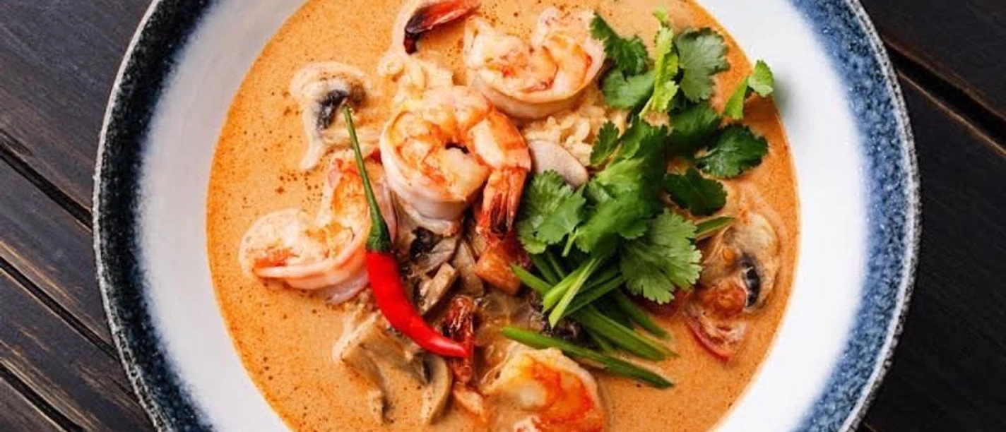 Chinglong Thai.jpeg Ein Teller mit thailändischer Garnelensuppe, garniert mit Koriander und Chilischote, auf Holztisch.