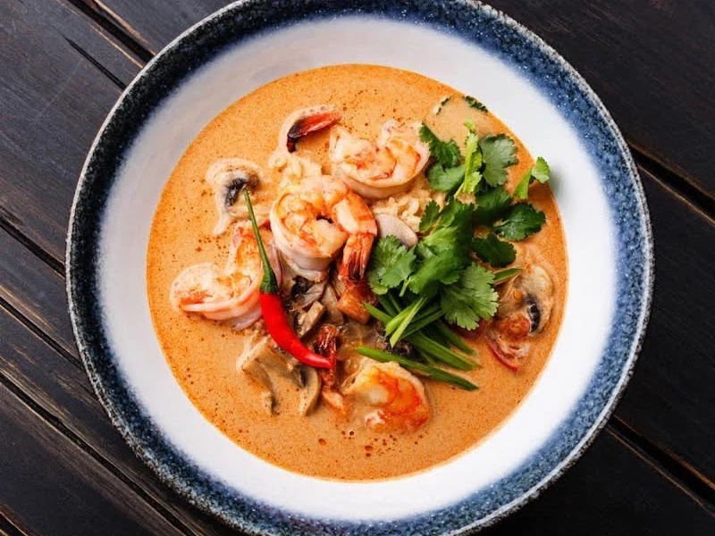 Chinglong Thai.jpeg Ein Teller mit thailändischer Garnelensuppe, garniert mit Koriander und Chilischote, auf Holztisch.