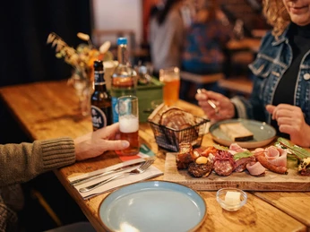 Johann Schäfer Brewery Ein gedeckter naturbelassender Holztisch gedeckt mit verschiedenen Gerichten. Eine Hand umschließt ein Bierglas und eine Frau schaut auf die servierten Speisen.A natural wooden table set with various dishes. A hand holds a beer glass and a woman looks at the food on the table.