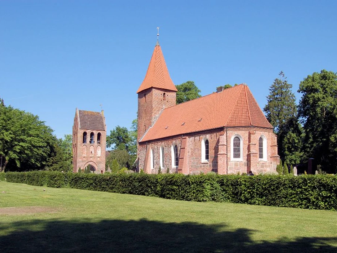 St. Ulrichs-Kirche - Seitenansicht mit Turm Backsteinkirche mit hohem Turm und rotem Ziegeldach, umgeben von grünen Bäumen und Rasenfläche.
