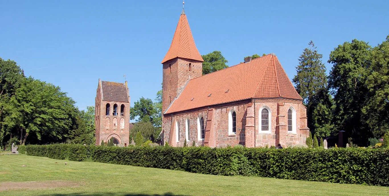 St. Ulrichs-Kirche - Seitenansicht mit Turm Backsteinkirche mit hohem Turm und rotem Ziegeldach, umgeben von grünen Bäumen und Rasenfläche.