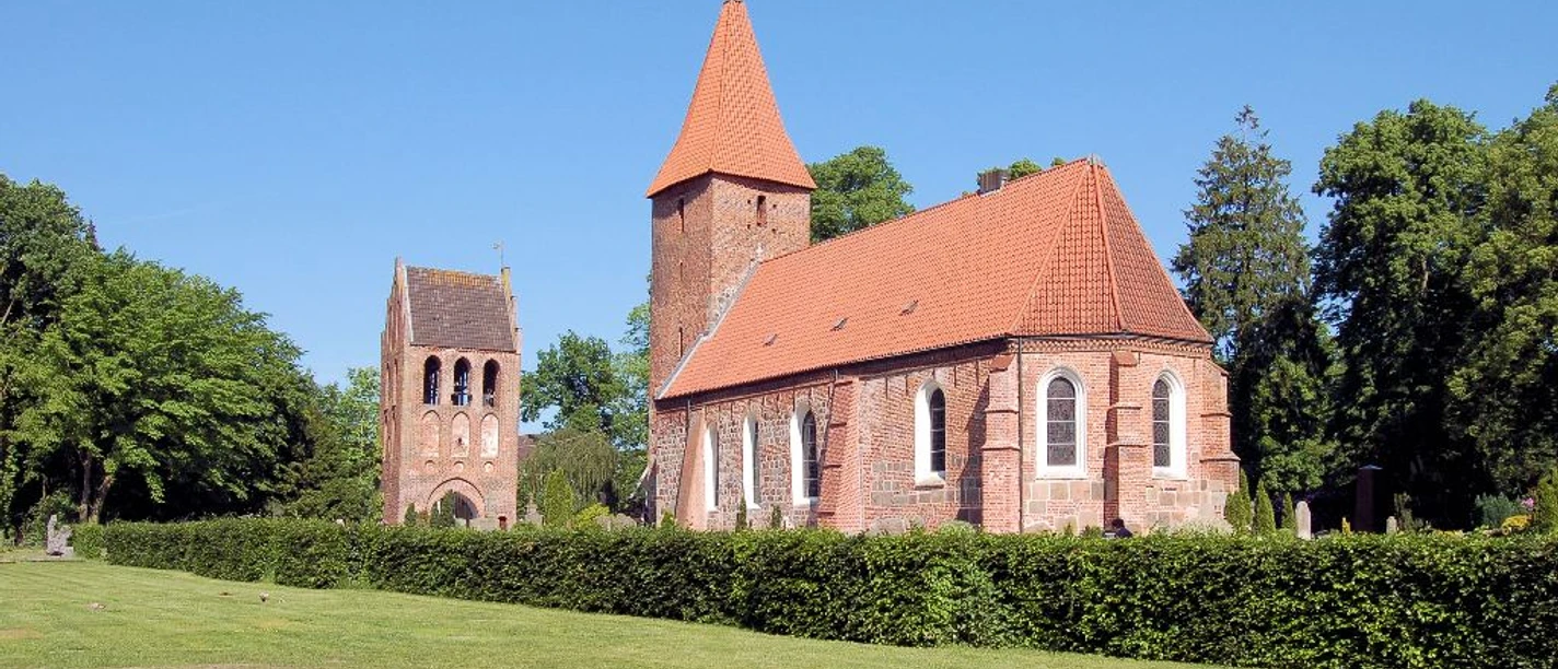 St. Ulrichs-Kirche - Seitenansicht mit Turm Backsteinkirche mit hohem Turm und rotem Ziegeldach, umgeben von grünen Bäumen und Rasenfläche.